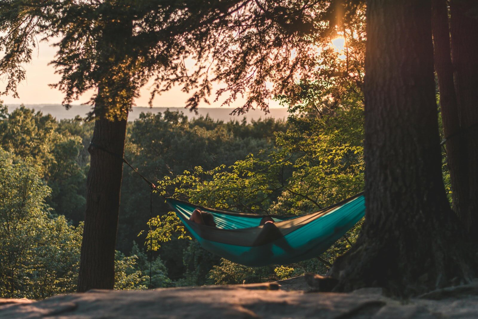 A scenic view of a forest with a hammock tied between two trees. The sun is setting in the background, casting a golden glow over the landscape, with a person relaxing inside the hammock